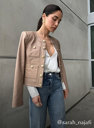 A woman stands against a gray concrete wall, wearing a beige jacket with gold buttons, a white blouse, and blue jeans. Her dark hair is pulled back, and she wears gold hoop earrings. Text at the bottom reads @sarah_najafi.