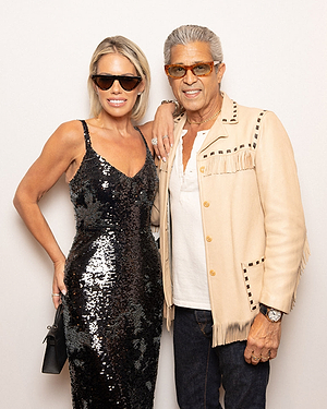 A woman in a black sequin dress and sunglasses stands next to a man in an off-white fringed jacket, white shirt, jeans, and orange-tinted glasses. Both are smiling and posing against a plain background.