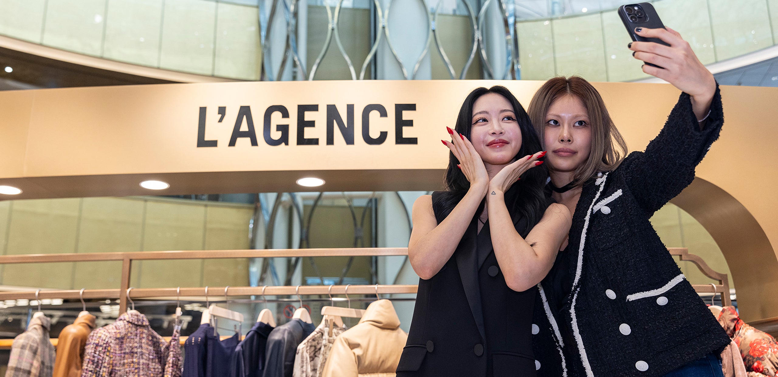 Two women pose together for a selfie in front of a clothing display and a large L’AGENCE sign in a modern retail store.