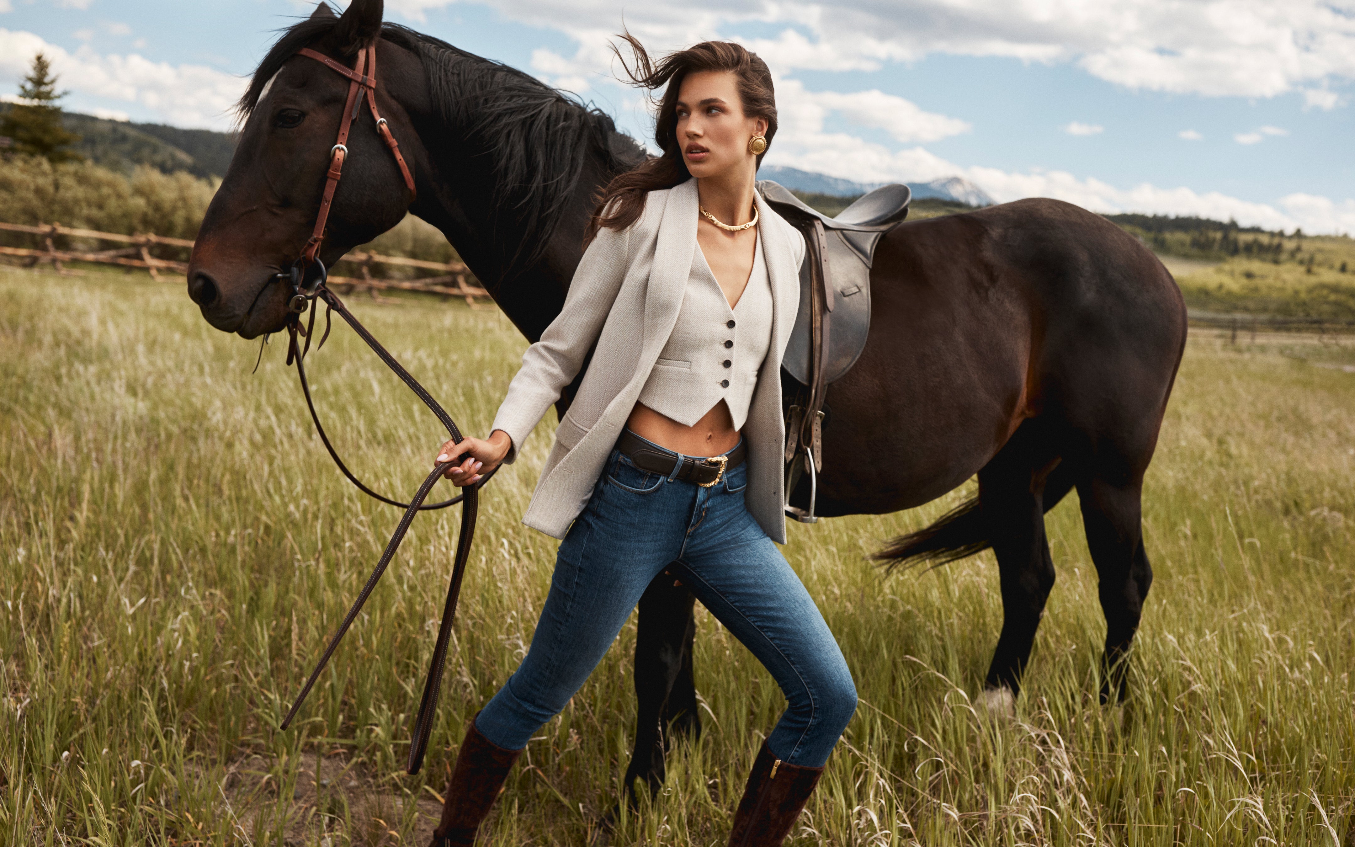 A woman in a blazer, vest, jeans, and boots stands confidently in a grassy field holding the reins of a brown horse. Mountains and a wooden fence are visible in the background under a partly cloudy sky.