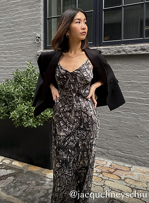 A woman stands outdoors against a gray brick wall, wearing a black-and-white patterned dress with a black blazer draped over her shoulders. She looks to the side with one hand on her hip. @jacquelineyschiu is written in the bottom right corner.