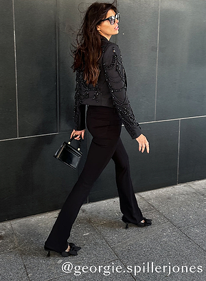 A woman with long brown hair, wearing sunglasses, a beaded black jacket, and black pants, walks on a sidewalk. She carries a small black handbag. The background is a dark tiled wall. Text reads @georgie.spillerjones.