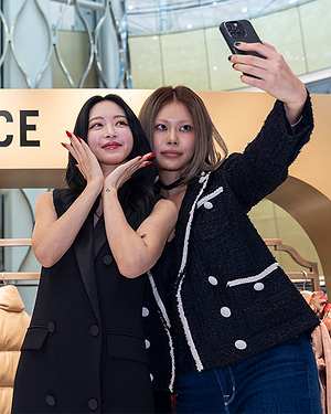 Two women pose closely together for a selfie in a stylish indoor setting; one woman smiles with her hands under her chin, while the other holds up a phone to take the picture.