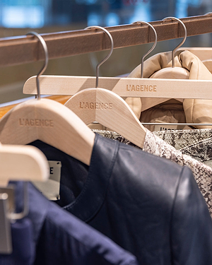 Four L’AGENCE wooden hangers hold assorted jackets and coats, including navy, patterned, and beige styles, neatly displayed on a clothing rack in a well-lit store.