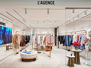 A stylish clothing boutique displays racks of women’s apparel, including dresses, jackets, and shirts. A round ottoman and small tables with flowers are in the center, with a large TV screen and modern decor throughout.