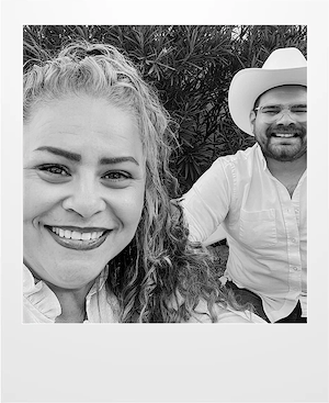 A smiling woman with curly hair takes a selfie with a man in a cowboy hat and glasses. Both are wearing light-colored shirts, and leafy bushes are in the background. The photo is in black and white.
