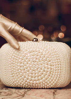 A gloved hand holds a small, elegant clutch purse decorated with intricate patterns of white pearls against a softly blurred background.