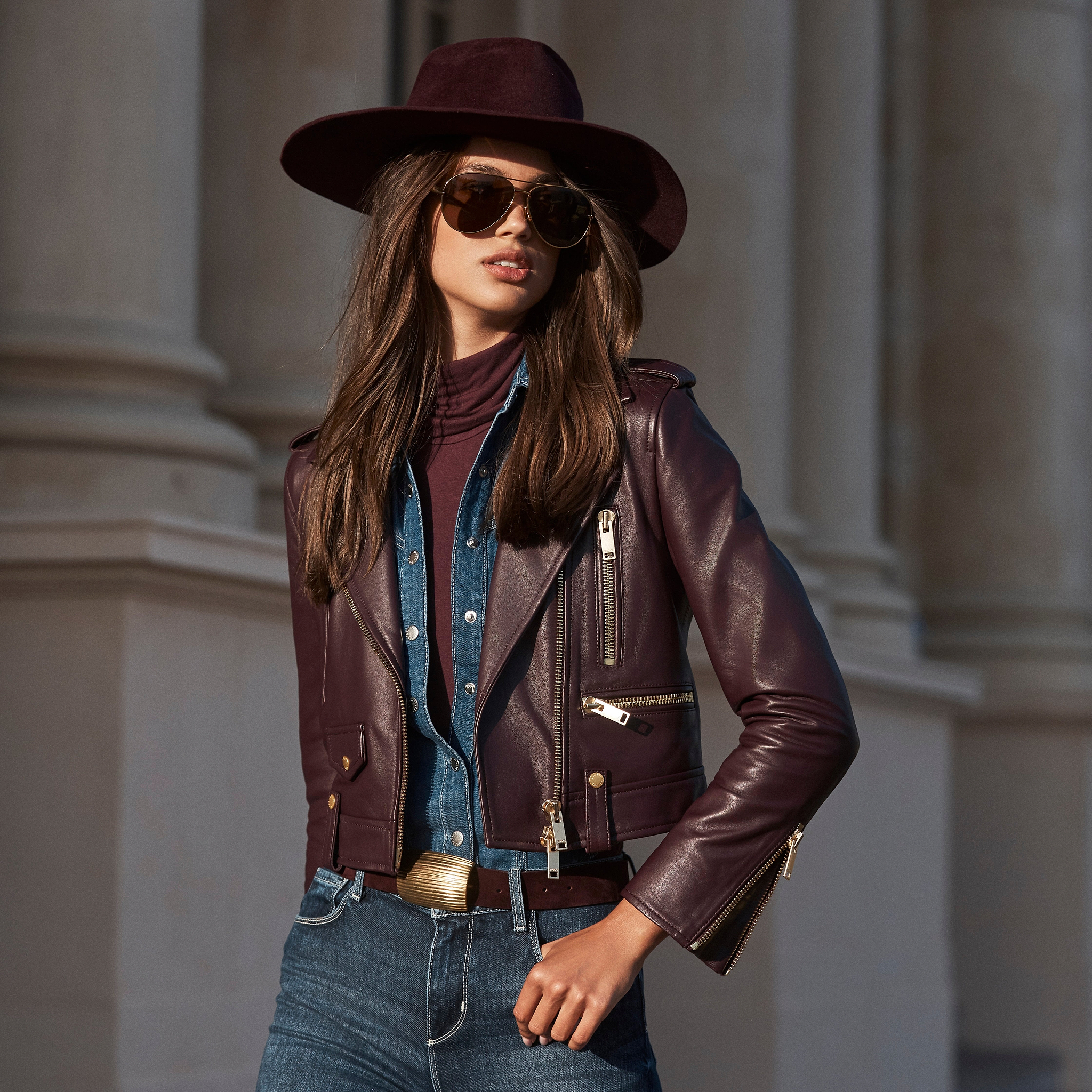 A woman wears a burgundy leather jacket over a denim shirt, a matching felt hat, and sunglasses.