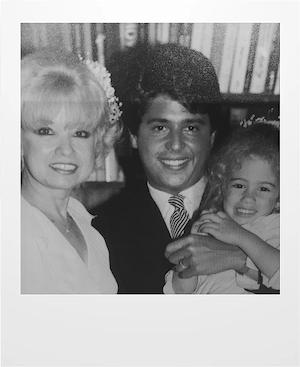 Black and white photo of two adults and a young child smiling together. The woman has light hair, the man has dark hair and wears a tie, and the child is hugging the man. Books are visible in the background.