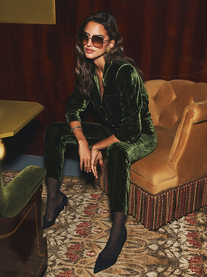 A woman with long brown hair and large sunglasses poses confidently on a gold couch, wearing the L'AGENCE Rebel Velvet Trouser in olive with black heels in a stylish, vintage room.