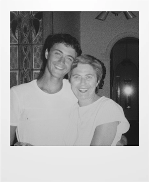 A young man and an older woman smile and embrace while standing indoors, captured in a black and white photograph. They are both wearing light-colored shirts. A decorative door and arched hallway are visible in the background.