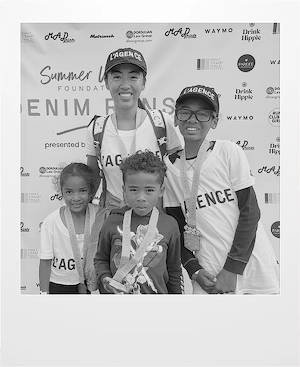 A smiling adult and three children wearing matching L’AGENCE shirts and medals pose together in front of a step-and-repeat event banner at the Denim Jeans Summer Fun Foundation event.