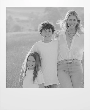 A woman stands outdoors with two smiling children, one boy and one girl, all dressed in light-colored casual clothes. They are standing close together in a sunlit field.