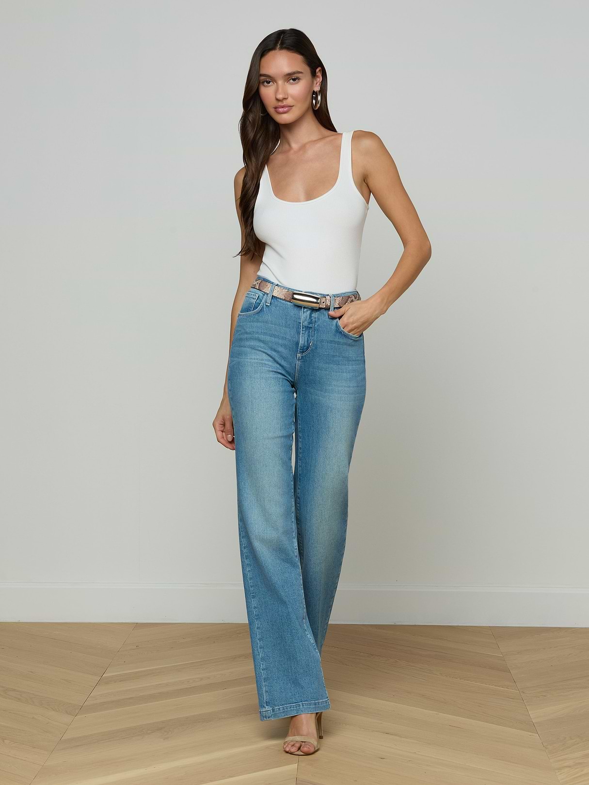 A woman stands against a plain white background wearing a white tank top, blue high-waisted wide-leg jeans, and heels, accessorized with the Marlowe Leather Belt in a python-embossed finish.