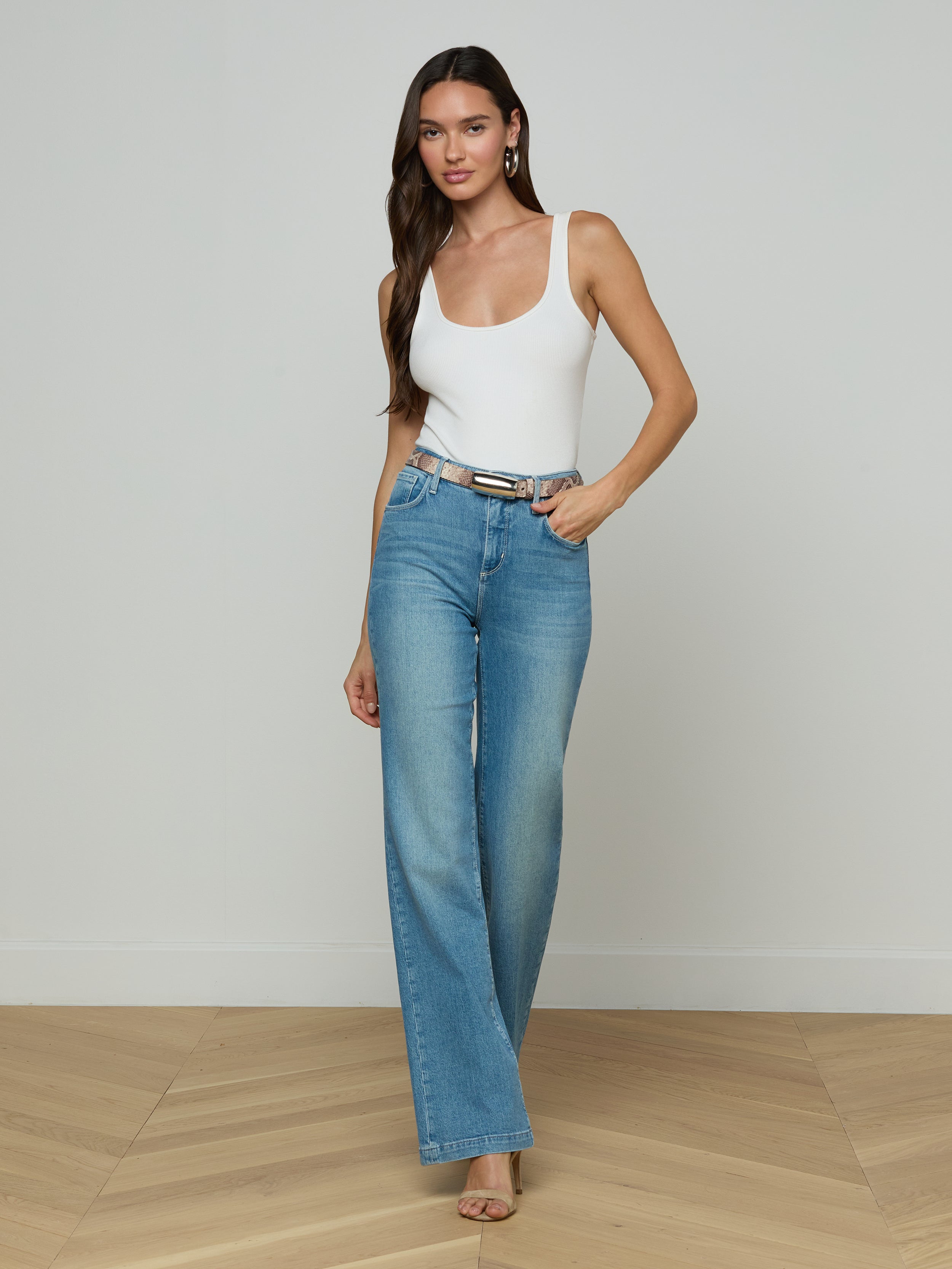 A woman stands against a plain white background wearing a white tank top, blue high-waisted wide-leg jeans, and heels, accessorized with the Marlowe Leather Belt in a python-embossed finish.