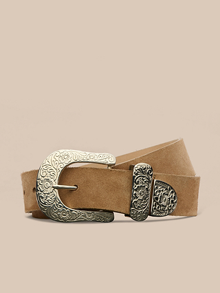 The Otera Suede Belt, a tan suede belt with an ornate silver buckle engraved with floral designs, is displayed coiled on a neutral beige background.