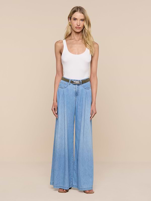 Une femme se tient debout devant un fond beige. Elle porte un haut blanc sans manches, un jean bleu clair à jambes larges, des sandales noires et la ceinture « Marlowe Suede » de couleur vert olive, dotée dune boucle plaquée argent qui apporte une touche minimaliste à sa tenue.