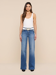 A woman stands facing forward, wearing a white tank top, blue high-waisted jeans, and black shoes. Her look is completed by the Tobar Leather Belt, featuring an embossed leather design and mixed-metal buckle, set against a plain white background.