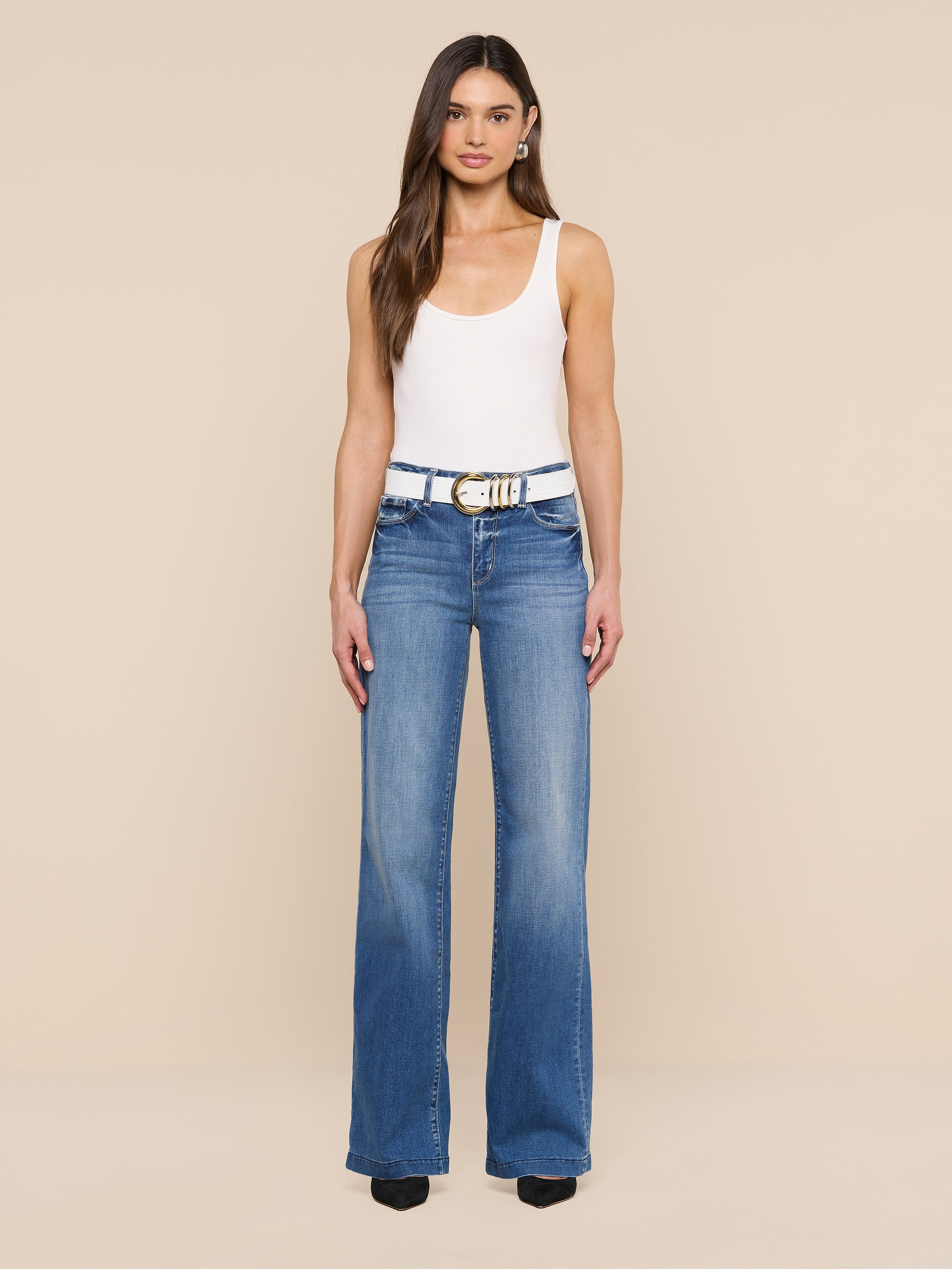 A woman stands facing forward, wearing a white tank top, blue high-waisted jeans, and black shoes. Her look is completed by the Tobar Leather Belt, featuring an embossed leather design and mixed-metal buckle, set against a plain white background.