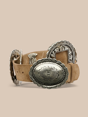 The Austin Suede Concho Belt features tan suede with large, ornate silver conchos that have engraved patterns and scalloped edges, shown on a plain beige background.