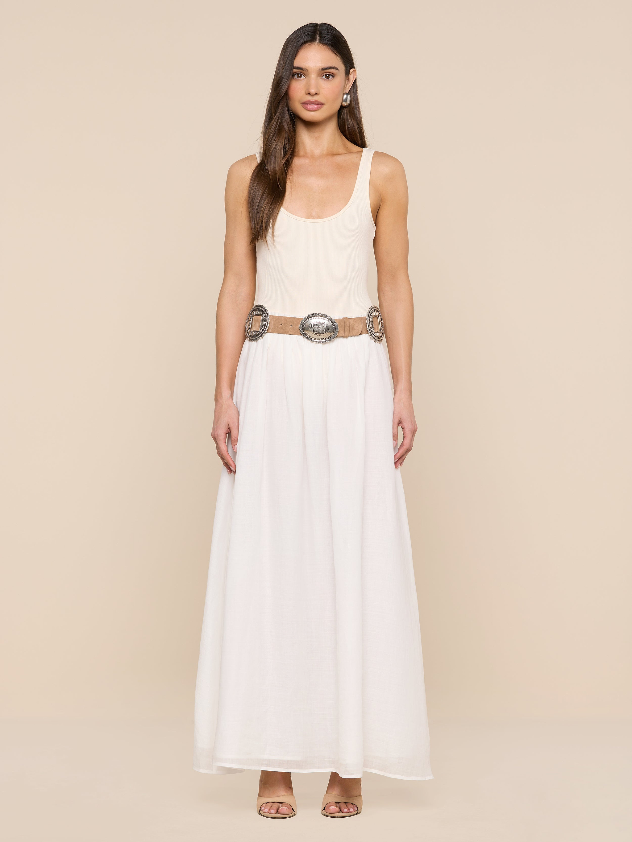 A woman stands against a beige background in a sleeveless cream top and long white skirt, styled with the Austin Suede Belt featuring large silver buckles. She has long hair and open-toed heels.
