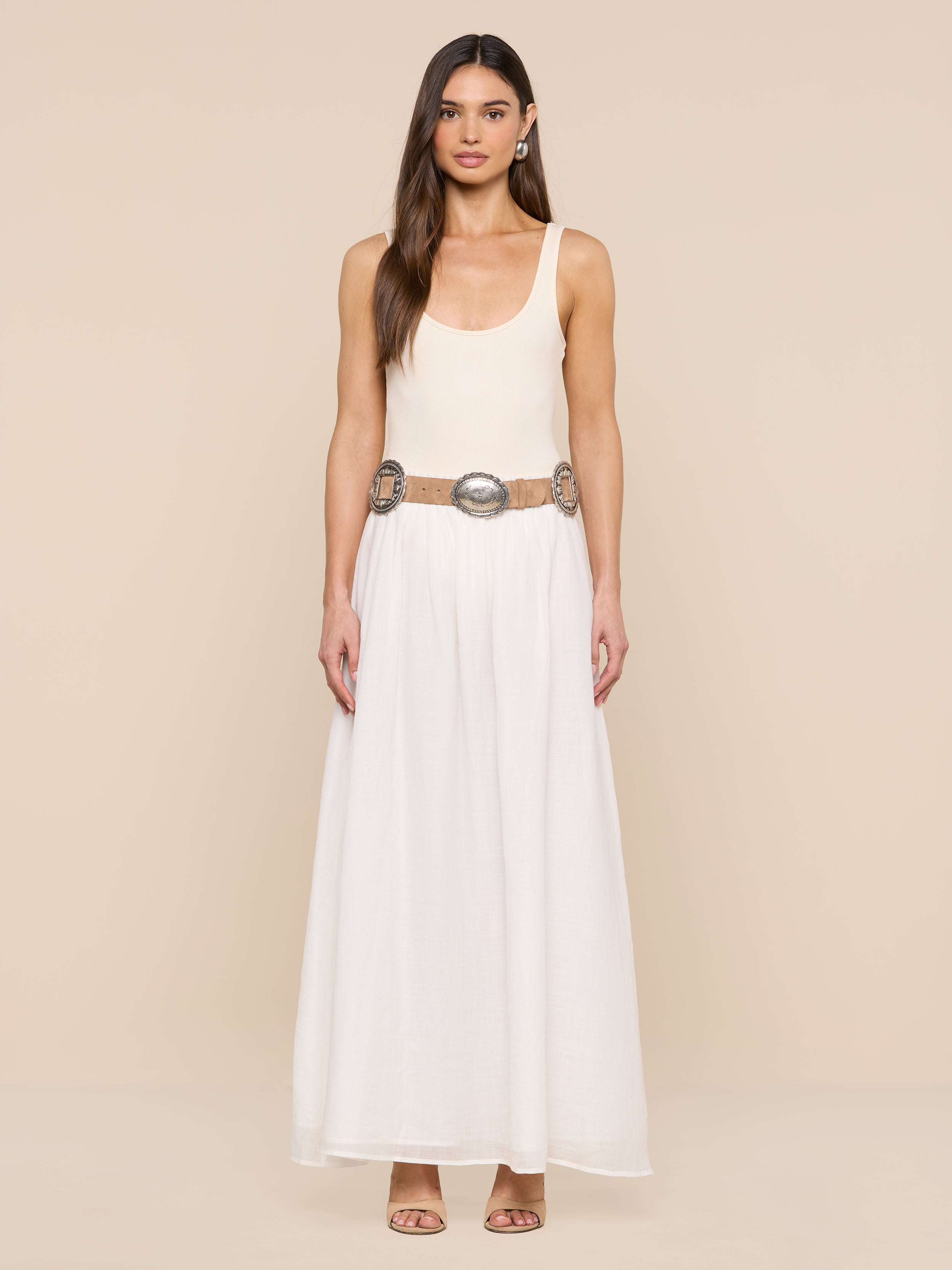 A woman stands against a beige background in a sleeveless cream top and long white skirt, styled with the Austin Suede Belt featuring large silver buckles. She has long hair and open-toed heels.