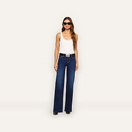 A woman with long brown hair poses against a white background, wearing dark sunglasses, a white tank top, the Solera Belt—a black calf hair belt with a silver buckle—and dark blue wide-leg jeans.