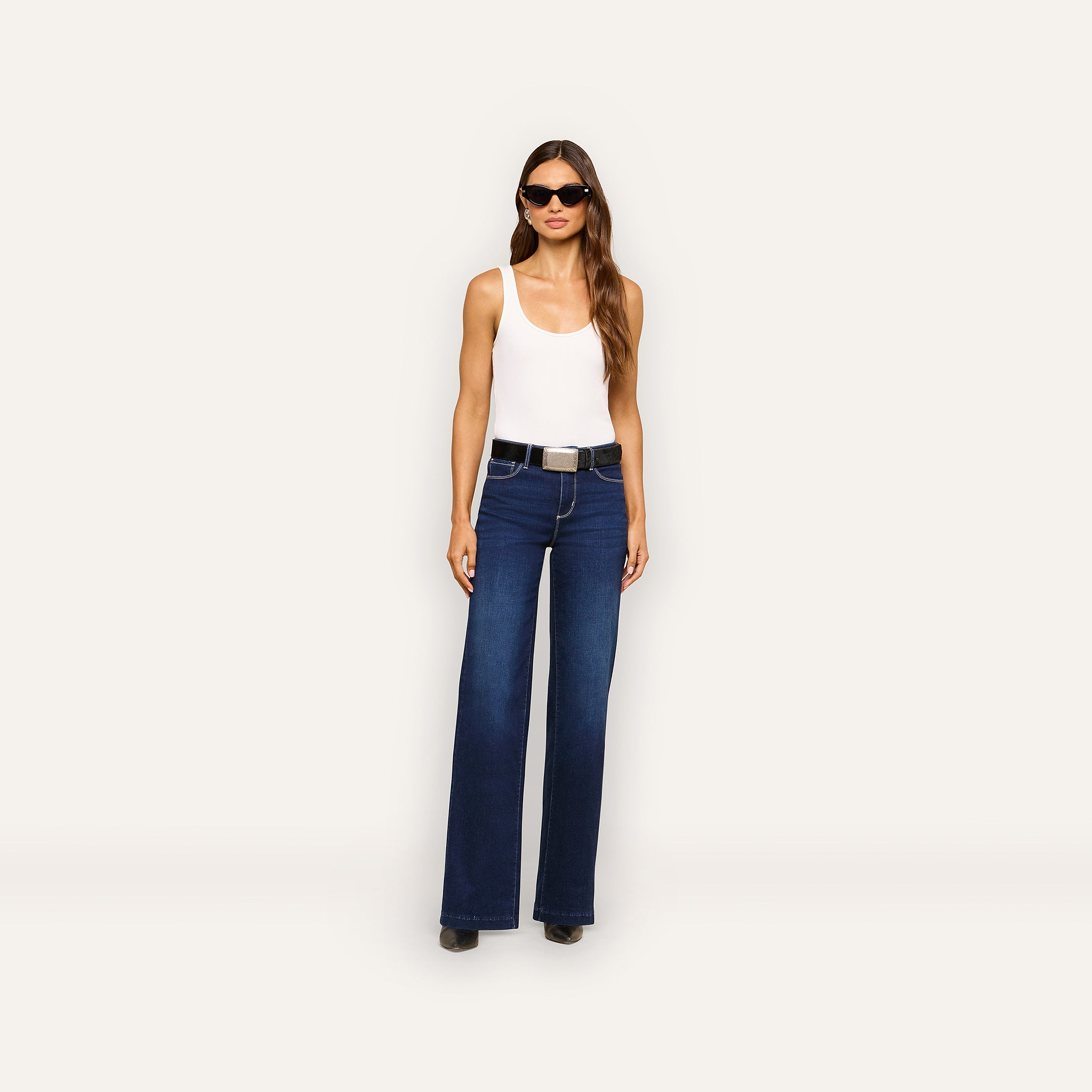 A woman with long brown hair poses against a white background, wearing dark sunglasses, a white tank top, the Solera Belt—a black calf hair belt with a silver buckle—and dark blue wide-leg jeans.