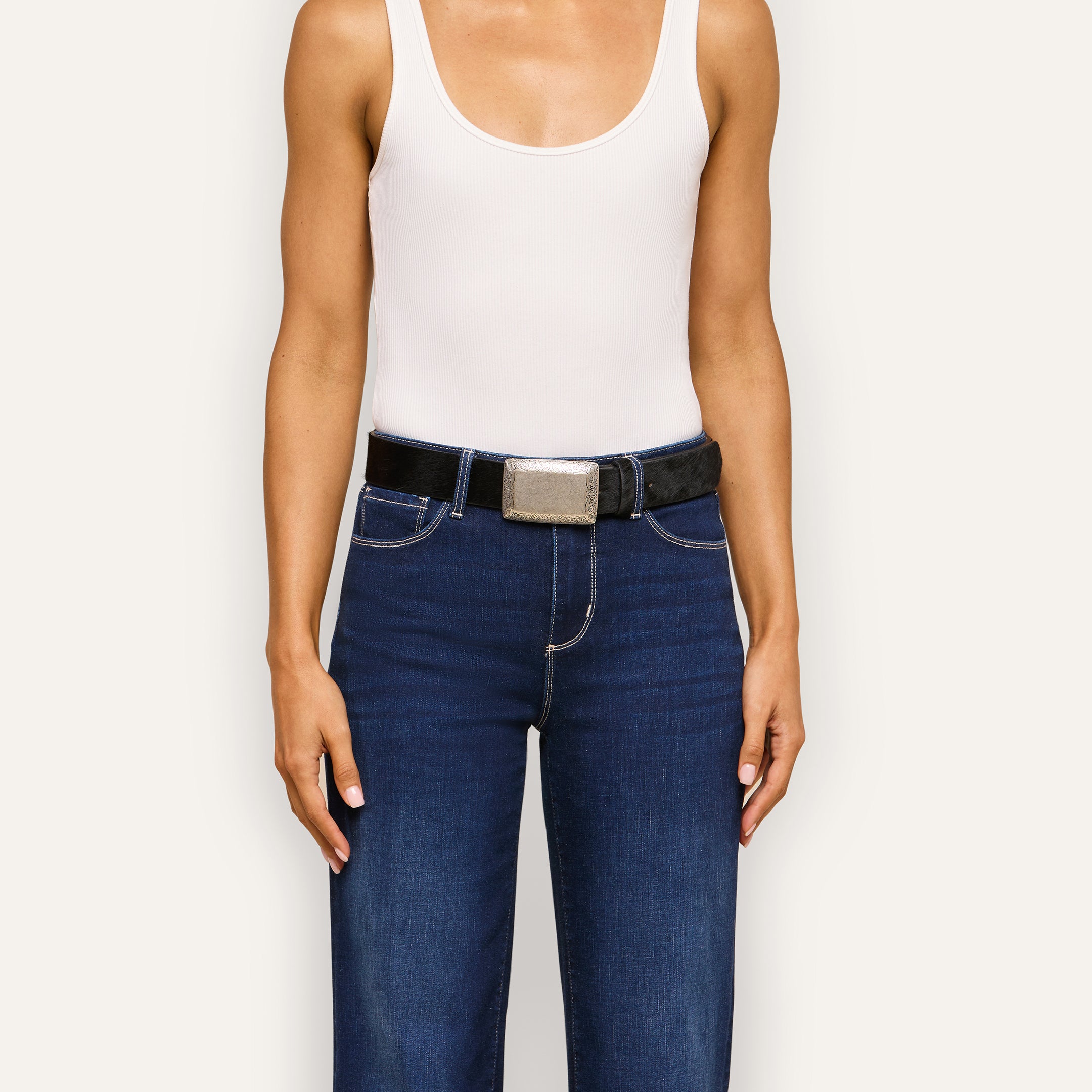 A person wearing a white sleeveless top, blue jeans, and the Solera Belt—handcrafted in LA from black calf hair with a large silver buckle—stands against a plain light background.