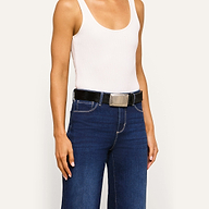 Person wearing a white tank top, blue jeans, and the Solera Belt—an Italian calf hair belt with a large gold-plated buckle—posing against a plain light background.