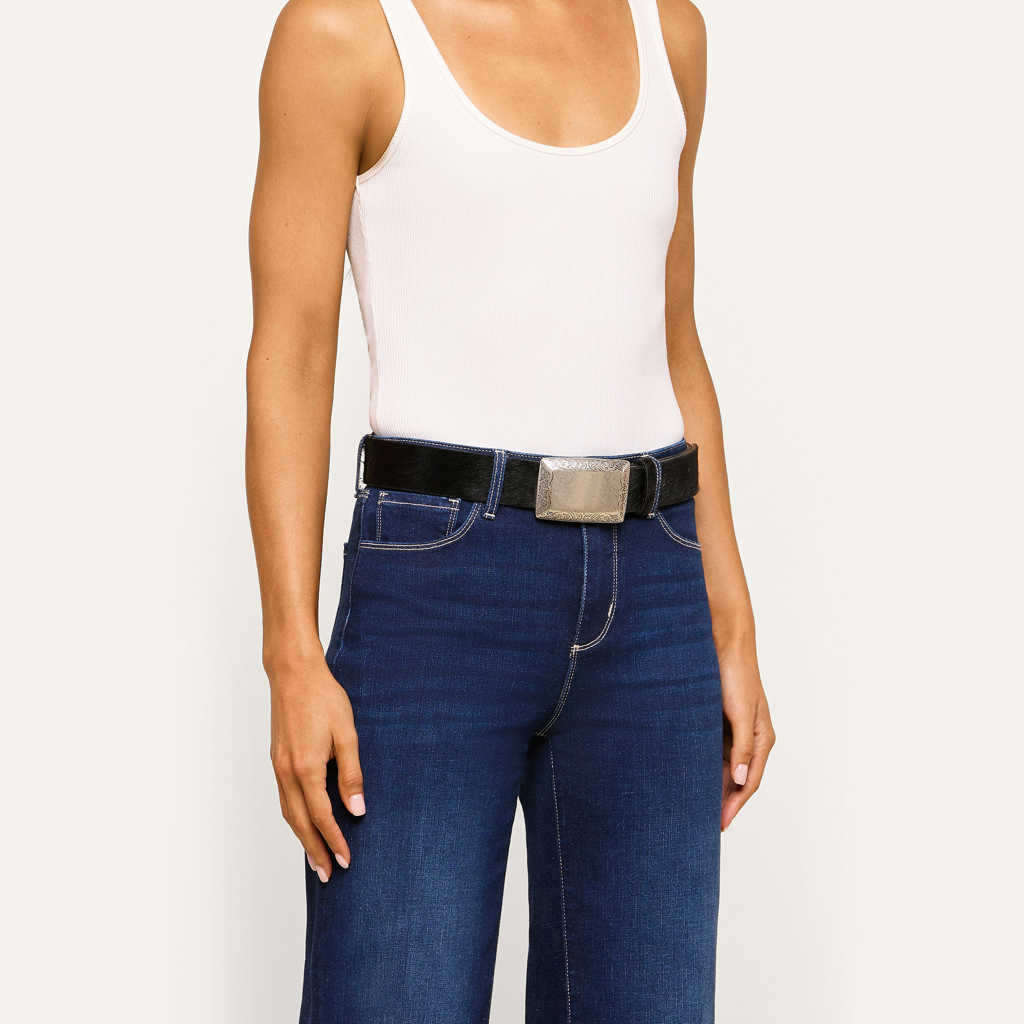 Person wearing a white tank top, blue jeans, and the Solera Belt—an Italian calf hair belt with a large gold-plated buckle—posing against a plain light background.