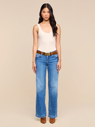 A woman stands facing forward against a plain beige background, wearing a beige tank top, blue high-waisted jeans cinched with the Sierra Belt—a handcrafted design featuring a brass buckle—and brown shoes.