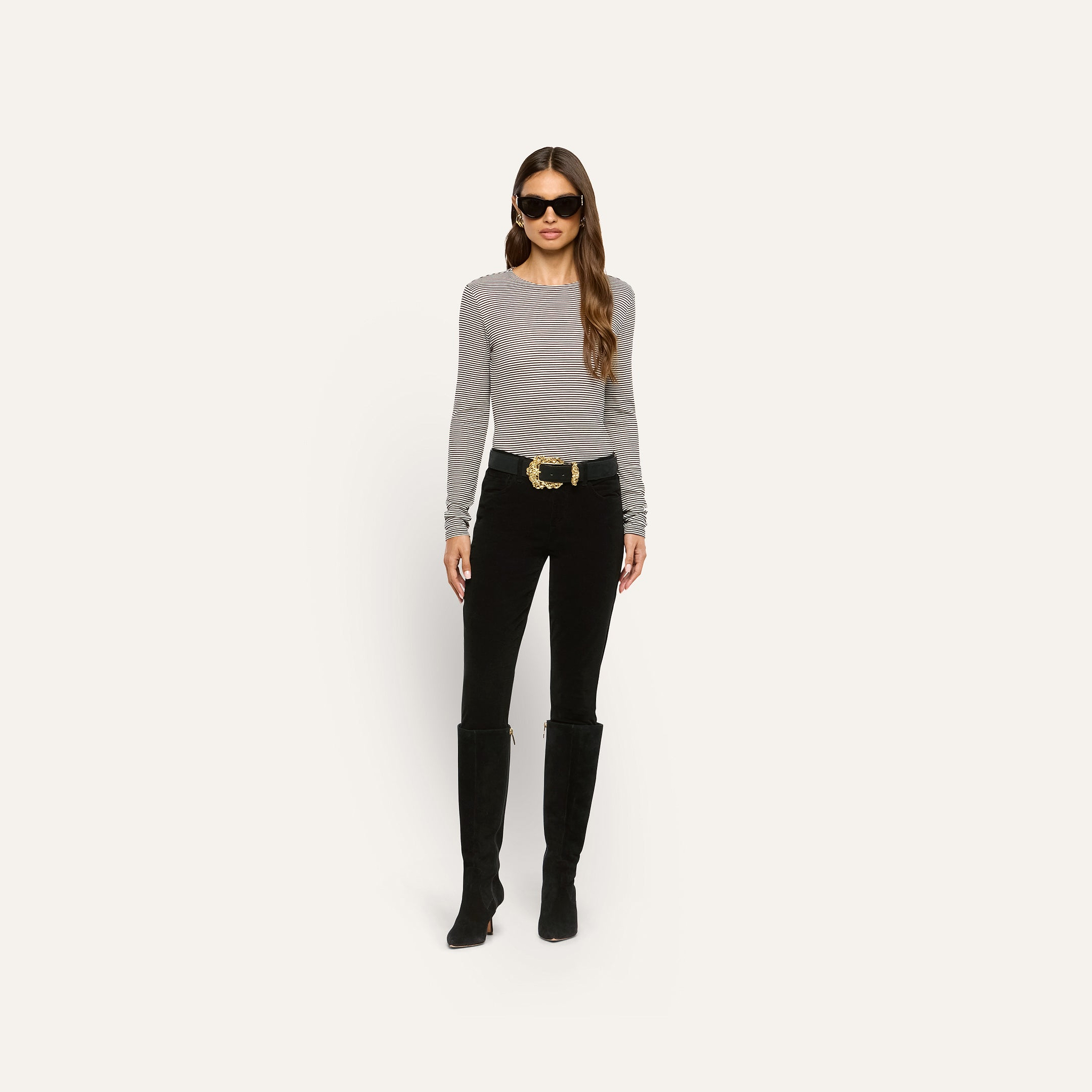 A woman wears a long-sleeved striped shirt, black pants, tall black boots, and a Caldera Suede Belt with a gold-plated buckle.