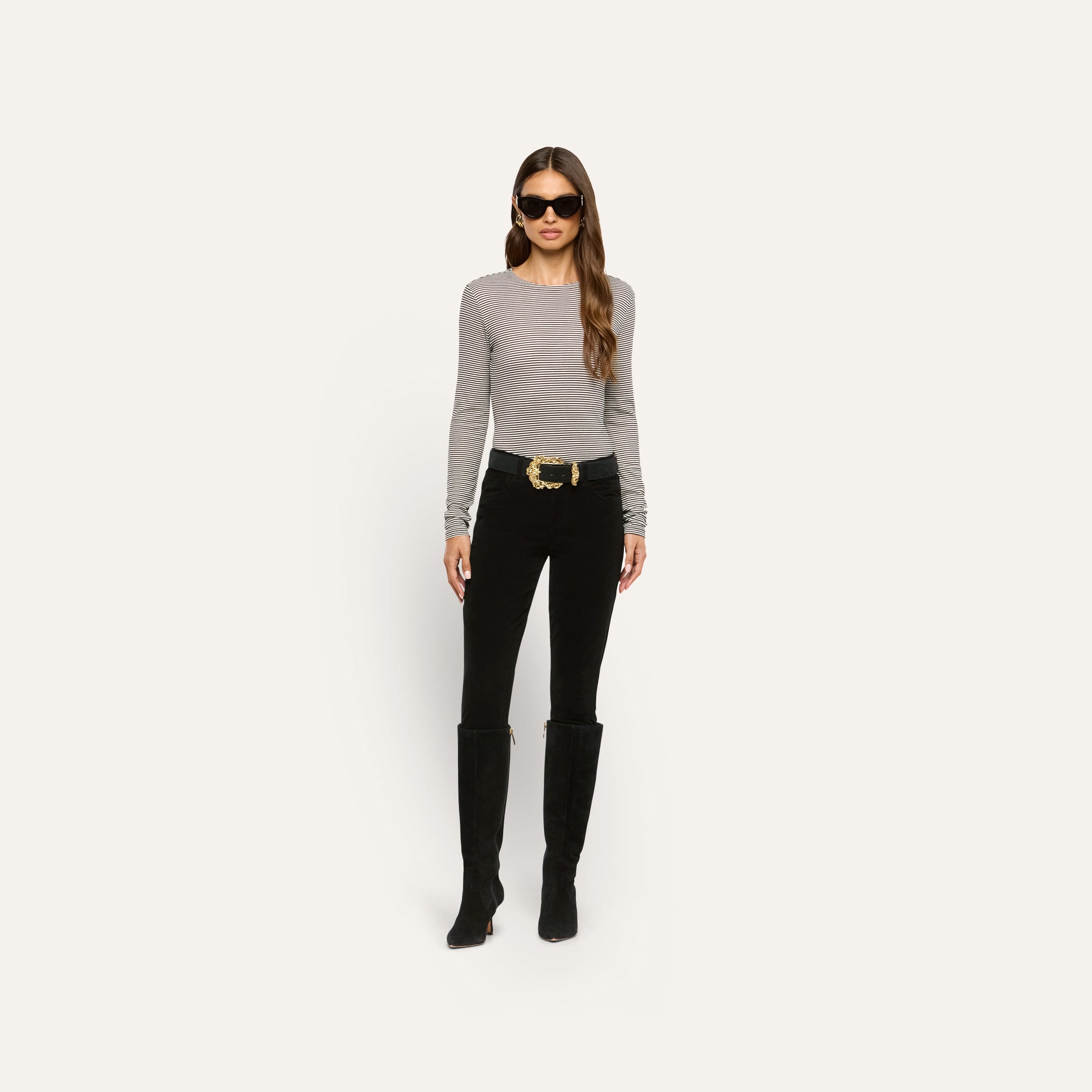 A woman wears a long-sleeved striped shirt, black pants, tall black boots, and a Caldera Suede Belt with a gold-plated buckle.