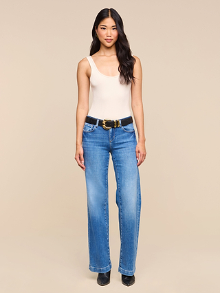 A woman stands against a beige background, wearing a sleeveless beige top, blue wide-leg jeans styled with the Yarros Leather Belt—a black suede belt featuring a gold-plated buckle—and black shoes. Her long, wavy hair frames her neutral expression.