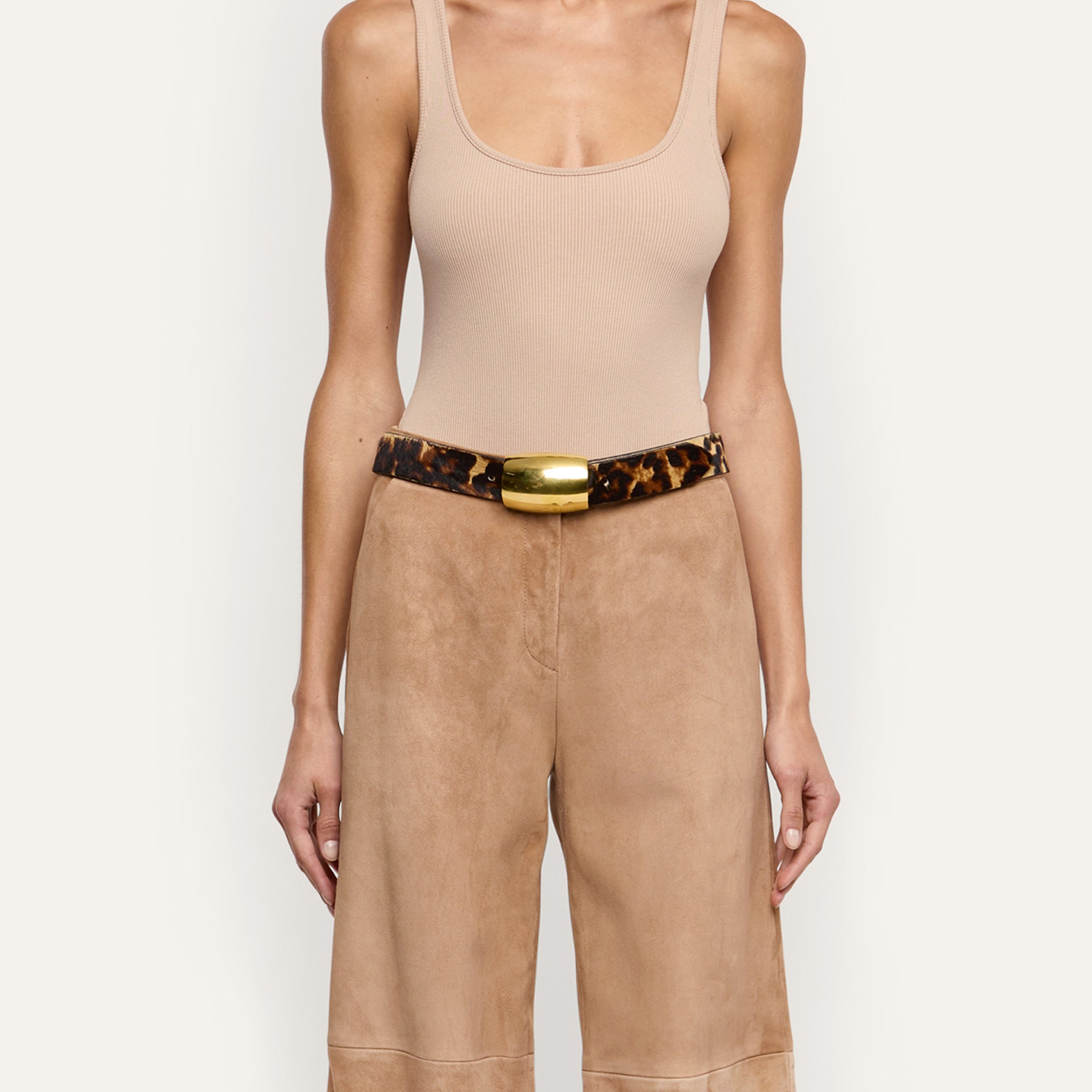 The Milou Belt, a handcrafted leopard print belt with a rectangular gold-tone metal buckle, worn over tan trousers and a beige ribbed tank top.