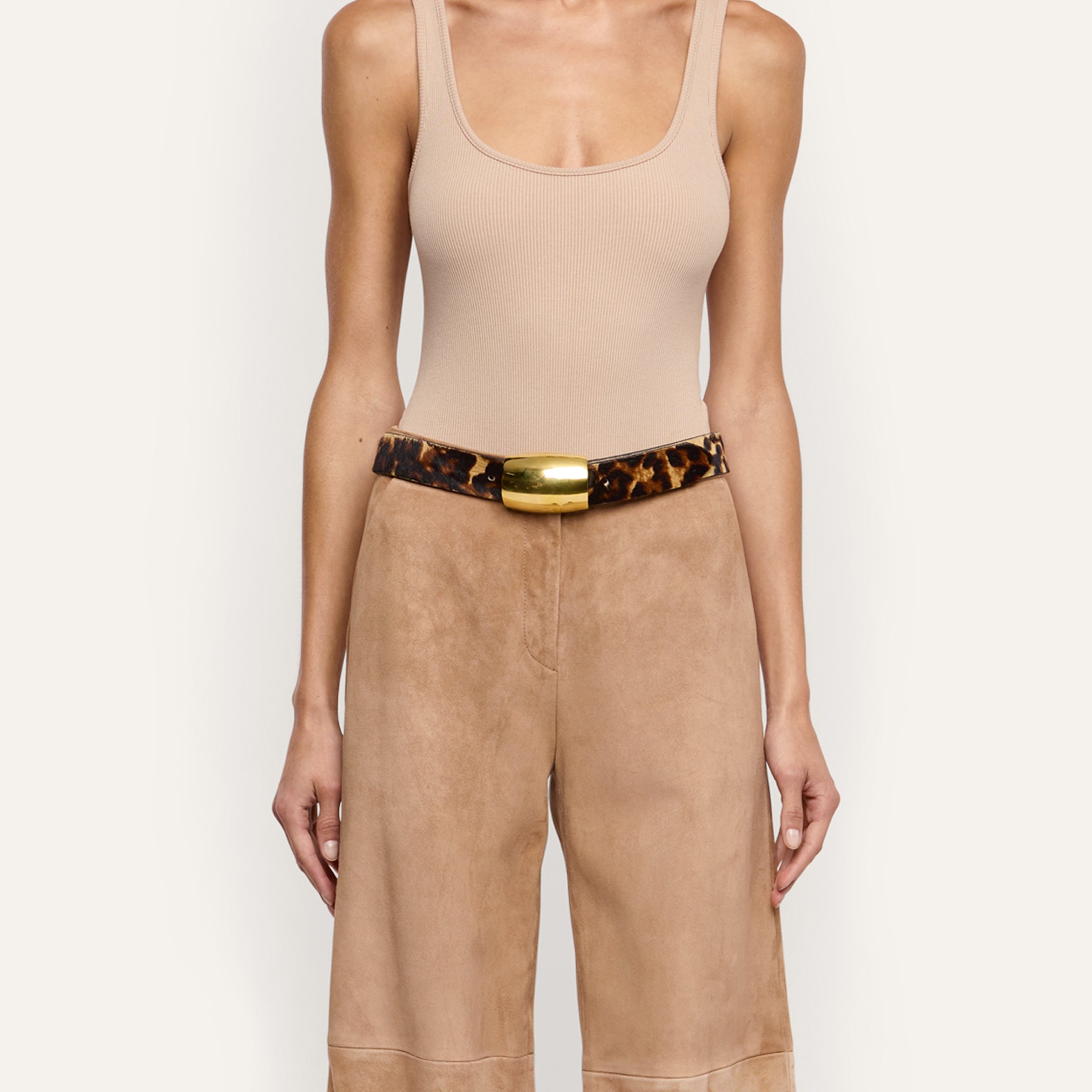 The Milou Belt, a handcrafted leopard print belt with a rectangular gold-tone metal buckle, worn over tan trousers and a beige ribbed tank top.