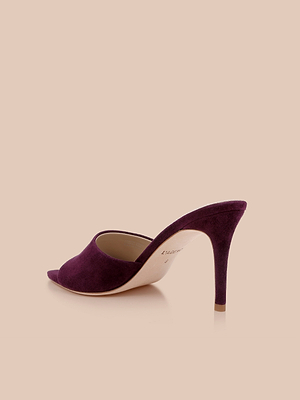 A Lolita Suede Open-Toe Mule in rich purple with a pointed toe and slim stiletto heel, showcased on a light beige background.