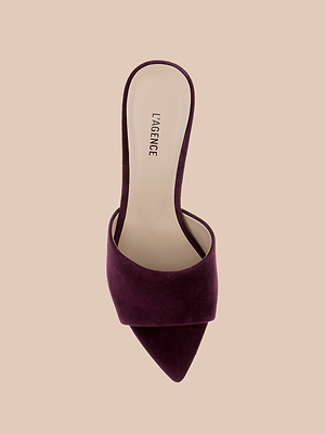 A single Lolita Suede Open-Toe Mule in dark burgundy with a pointed toe, wide upper strap, and open back is shown from above on a beige background. The insole features the L’AGENCE label.