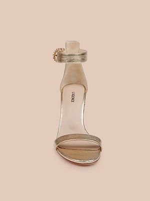 Il sandalo Gisele in pelle è caratterizzato da pelle dorata, cinturino singolo in punta, cinturino alla caviglia con catena dorata e soletta beige con letichetta LAURENCE, una scelta elegante per le invitate al matrimonio, su uno sfondo color pesca pallido.