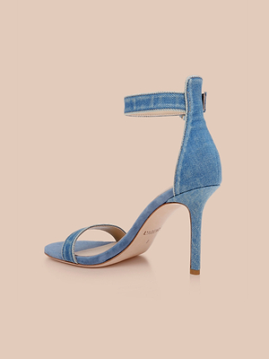 La sandalia Gisele Denim presenta una parte superior de tela vaquera azul, correa en el tobillo, tacón alto y puntera abierta, sobre un fondo melocotón claro.