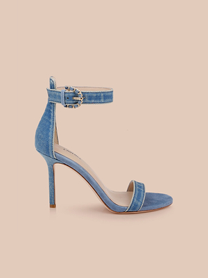 La sandalia Gisele Denim presenta una parte superior vaquera, correa de tobillo con una elegante hebilla, puntera abierta y tacón de aguja, todo ello sobre un fondo melocotón claro.