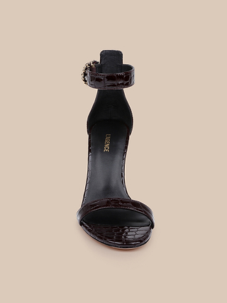 Front view of a dark brown, crocodile embossed leather, open-toe Gisele sandal with an ankle strap and gold buckle, displayed on a beige background.