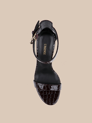 A single black, crocodile-embossed leather heeled sandal featuring an ankle strap and buckle, displayed from above on a beige background. The insole displays LAGENCE in gold lettering, showcasing the sleek sophistication of the Gisele Leather Sandal.