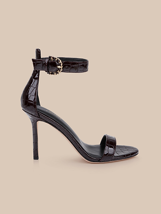 The Gisele Leather Sandal, crafted from crocodile-embossed leather in dark brown, features a high heel, an ankle strap with a decorative gold buckle, and is displayed on a light beige background.