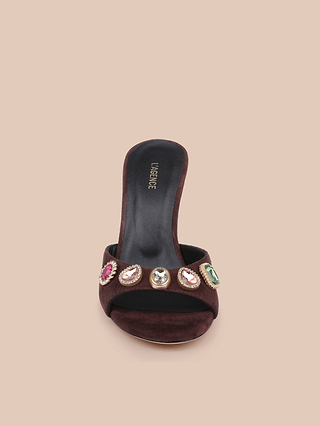 The Avery Suede Open-Toe Mule is a black heeled sandal featuring a wide strap embellished with large, colorful gemstones, displayed against a light beige background. This jeweled sandal brings a glamorous accent to any outfit.