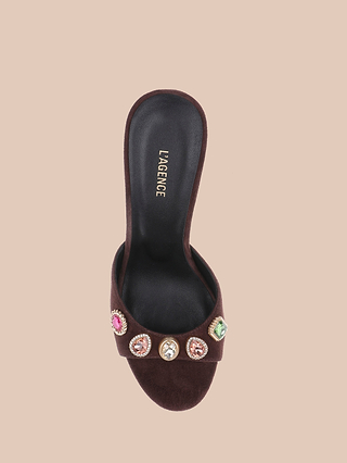 The Avery Suede Open-Toe Mule is a mahogany brown sandal with a black insole labeled LAGENCE, adorned with colorful gem embellishments on the front strap and showcased against a beige background.