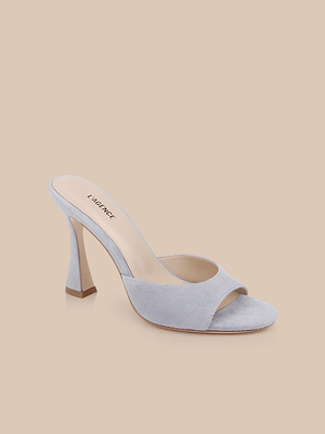The Avery Suede Open-Toe Mule is shown in pale blue suede with a sculpted high heel and open toe, set against a plain beige background. The memory foam insole ensures comfort and features the LAGENCE brand name.