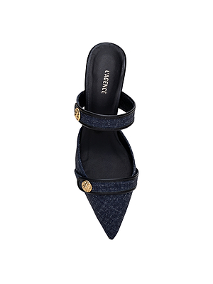 The Gilliane Heeled Mule by L’AGENCE features a pointed-toe, open-back denim design with two foot straps embellished with gold lion-embossed buttons and a black insole bearing the brand name, offering vintage-inspired elegance.