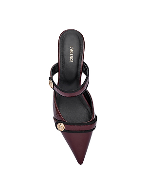 A top view of the Gilliane Heeled Mule by L'AGENCE in wine patent leather shows two straps with lion-embossed buttons and a pointed toe. The heeled design blends vintage and contemporary style with a black insole labeled 